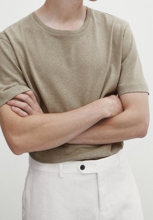Personne portant une chemise beige à manches courtes et un pantalon blanc, debout les bras croisés devant un fond clair uni.