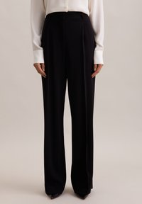 Czarne szerokie spodnie wykonane z gładkiego materiału, z wysokim stanem, plisami z przodu i dopasowanym krojem. Zestawione z białą bluzką.