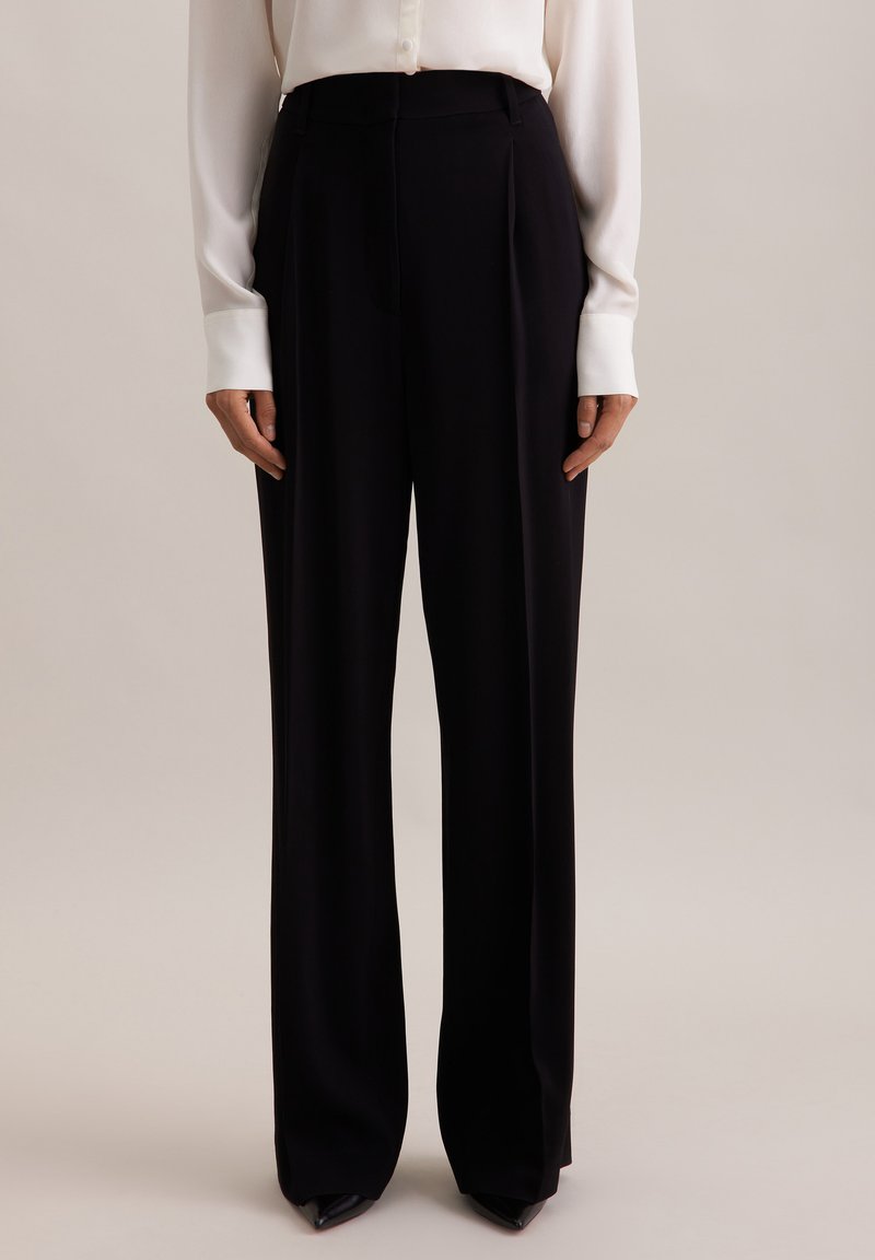 Czarne szerokie spodnie wykonane z gładkiego materiału, z wysokim stanem, plisami z przodu i dopasowanym krojem. Zestawione z białą bluzką.