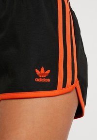 Black athletic shorts with orange trim and three stripes. Features an orange adidas logo on the lower left side. Textured material.
