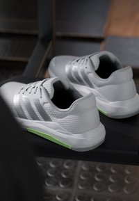 Paire de chaussures de sport blanches avec semelles vert clair posées sur un banc noir à l'intérieur avec un sol carrelé visible en dessous.