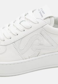 Baskets en cuir blanc avec un logo texturé sur le côté, lacets plats blancs et semelle en caoutchouc lisse. Design épuré avec des détails de couture subtils.