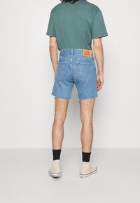 Pantalones cortos de mezclilla en azul claro con dobladillo deshilachado. Presenta dos bolsillos traseros, etiqueta de Levi's, combinados con una camiseta verde y zapatillas beige.