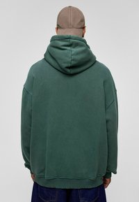 Groene oversized hoodie gemaakt van zachte, gestructureerde stof, met een grote capuchon en ribgebreide zoom. Gepaarde met donkerblauwe spijkerbroek.