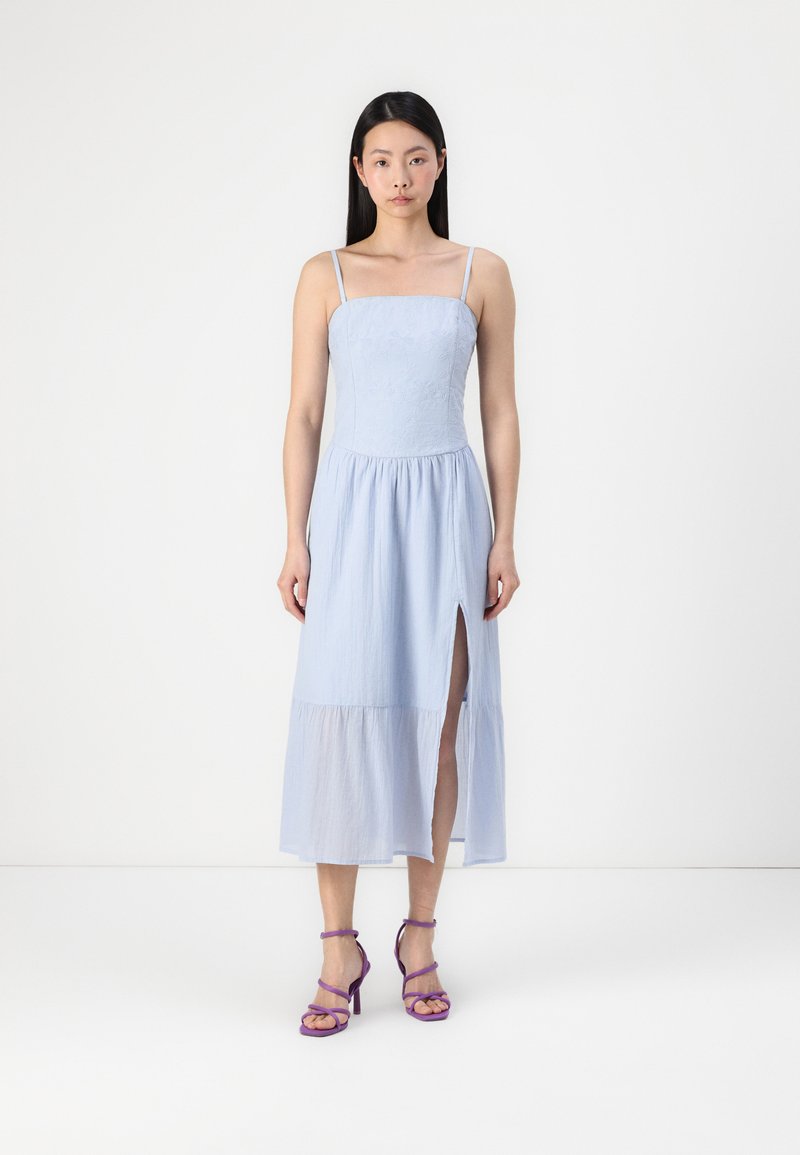 Rochie maxi fără mâneci, de culoare albastru deschis, cu un corset ajustat, textură florală și fante laterale. Purtată cu tocuri mov cu bretele.