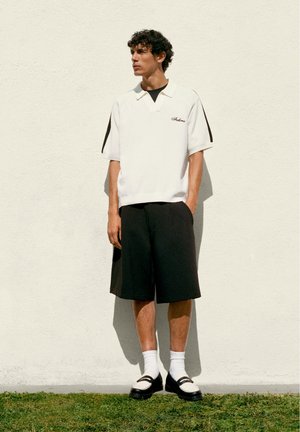 Junger Mann trägt weißes kurzärmeliges Polo, schwarze Shorts, weiße Socken und schwarz-weiße Loafers, steht an einer weißen Wand auf Gras.