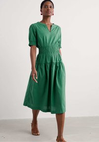 Grön klänning med kort ärm och V-ringning, smockad midja och tiered kjol. Tillverkad av lättviktig tyg med en mjuk textur.