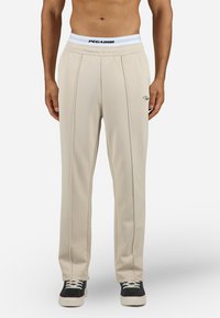 Beige treningsbukser med en myk tekstur, elastisk midje med en hvit logo, og sidesømmer. Kombinert med svarte joggesko.