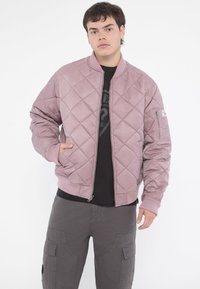 Quilted bomber jacket in light mauve, featuring a ribbed collar and cuffs, zippered front, and side pockets. Worn over a black t-shirt.