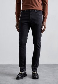 Svarta jeans i slim fit med fem fickor och traditionell knäppning, matchade med svarta loafers och en brun tröja.