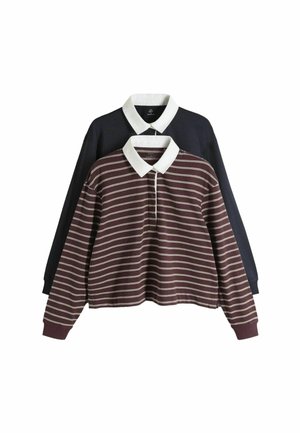 Mladá žena s krátkymi vlasmi nosiaca bordovo-biele pásikavé rugby tričko a svetlo modré džínsy s rukami v vreckách.