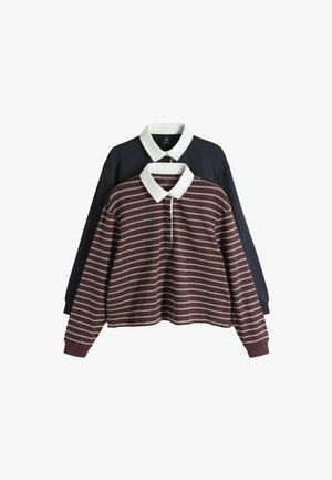 Fiatal nő rövid hajjal, bordó-fehér csíkos rögbi pólóban és világoskék farmerral, kezét a zsebében tartva.