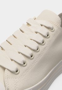 Κοντινή λήψη ενός πάνινου αθλητικού παπουτσιού σε κρεμ χρώμα με φαρδιές, επίπεδες κορδόνια περασμένα μέσα από μεταλλικά περβάζια και ραφές διακόσμησης στο πάνω μέρος.