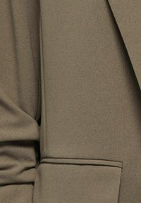Gros plan d'une poche de veste en tissu marron avec des coutures visibles et une partie de la manche et des coutures du corps.