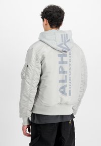 Světle šedá bomber bunda s texturovaným povrchem, opatřená kapucí, elastickými manžetami a výrazným logem na zadní straně.