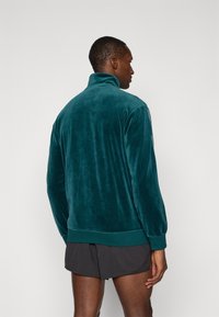 Zöld velúr pulóver magas gallérral, hosszú ujjakkal és bordázott aljjal. Puha textúra, minimális varrásokkal a letisztult sziluett érdekében.