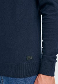 Marineblauer Strickpullover aus Wollmischung, mit gerippten Bündchen und Saum, sowie einem kleinen Markenlabel, das am unteren Ende angenäht ist.
