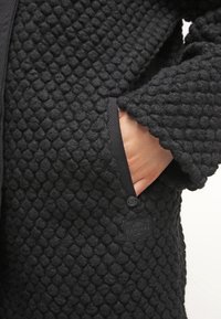 El abrigo negro de textura presenta una tela con un patrón de diamantes, un bolsillo frontal y una etiqueta cosida en el dobladillo, con una apariencia suave y cálida.
