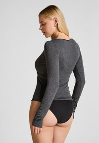 Vrouw in een aansluitende grijze top met lange mouwen en zwarte hooggesneden onderbroek, staande in profiel met het hoofd iets naar beneden gekanteld.