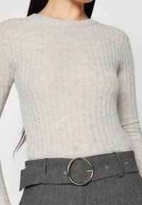 Donna che indossa un maglione a costine grigio chiaro infilato in pantaloni a spina di pesce grigio scuro, con una cintura larga dotata di una fibbia rotonda in argento.