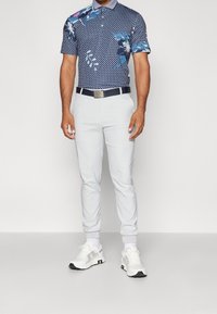 Polo azul marino con estampado floral y patrón geométrico, combinado con pantalones gris claro y zapatillas blancas. Incluye un cinturón oscuro y puños con ribete.