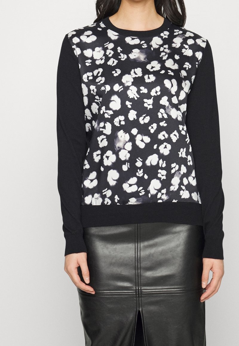 Žena v černém topu s dlouhým rukávem s bílými květinovými vzory a černé kožené sukni s předním rozparkem.