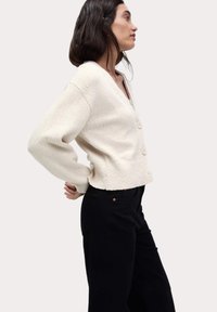 Cardigan en tricot couleur crème avec un col en V, doté de trois boutons sur le devant et d'une coupe courte. Assorti à un pantalon noir à jambes larges.