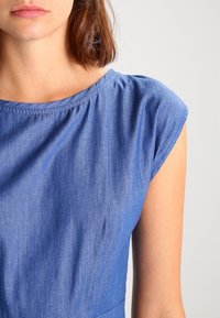 Jeansklänning med rund halsringning och korta ärmar. Tyget är ljusblått och något texturerat, med subtila rynkor vid axlarna.