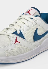 Λευκό και μπλε αθλητικό παπούτσι Nike Air Jordan με κόκκινα λογότυπα Jumpman και λευκά κορδόνια σε απλό φωτεινό φόντο.