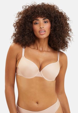 Mujer con cabello rizado que lleva un sujetador color nude y ropa interior a juego, de pie sobre un fondo blanco liso.