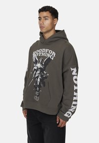 Hoodie vert olive avec un imprimé graphique d'un ange tenant une épée, doté d'une poche frontale et de texte blanc sur les manches.