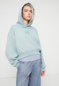 Svetlo modrá skrátená mikina s predným vreckom, s logom "GAP" v tmavšej farbe. Kombinovaná s modrými džínsami s vysokým pásom.