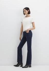 Blue denim flared jeans with front pockets, silver hardware, and a dark wash. Paired with a fitted white ribbed short-sleeve top.