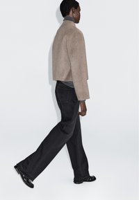 Beżowa krótka kurtka z mieszanki wełny, szary golf, czarne spodnie z szerokimi nogawkami i czarne błyszczące buty. Gładkie tło, widok z boku modelki idącej.