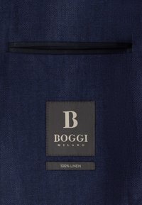 Tissu en chevrons bleu foncé avec une poche passepoilée noire et une étiquette noire portant les inscriptions "B BOGGI MILANO" et "100% LIN" en dessous.