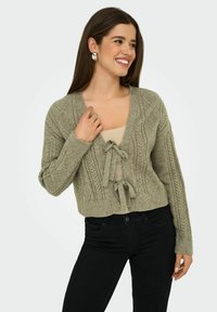 Cardigan cropped en tricot torsadé vert olive avec décolleté en v et fermeture à nouer, présentant des motifs texturés et des poignets côtelés, associé à un haut beige.