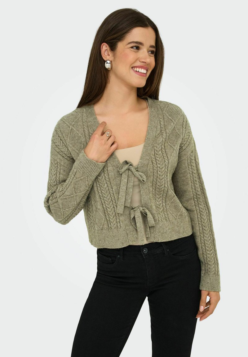 Cardigan cropped en tricot torsadé vert olive avec décolleté en v et fermeture à nouer, présentant des motifs texturés et des poignets côtelés, associé à un haut beige.