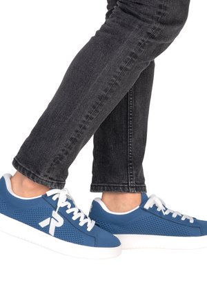 Pieds portant des baskets en mesh bleu avec des lacets et semelles blanches, assorties à un jean en denim noir sur fond blanc.