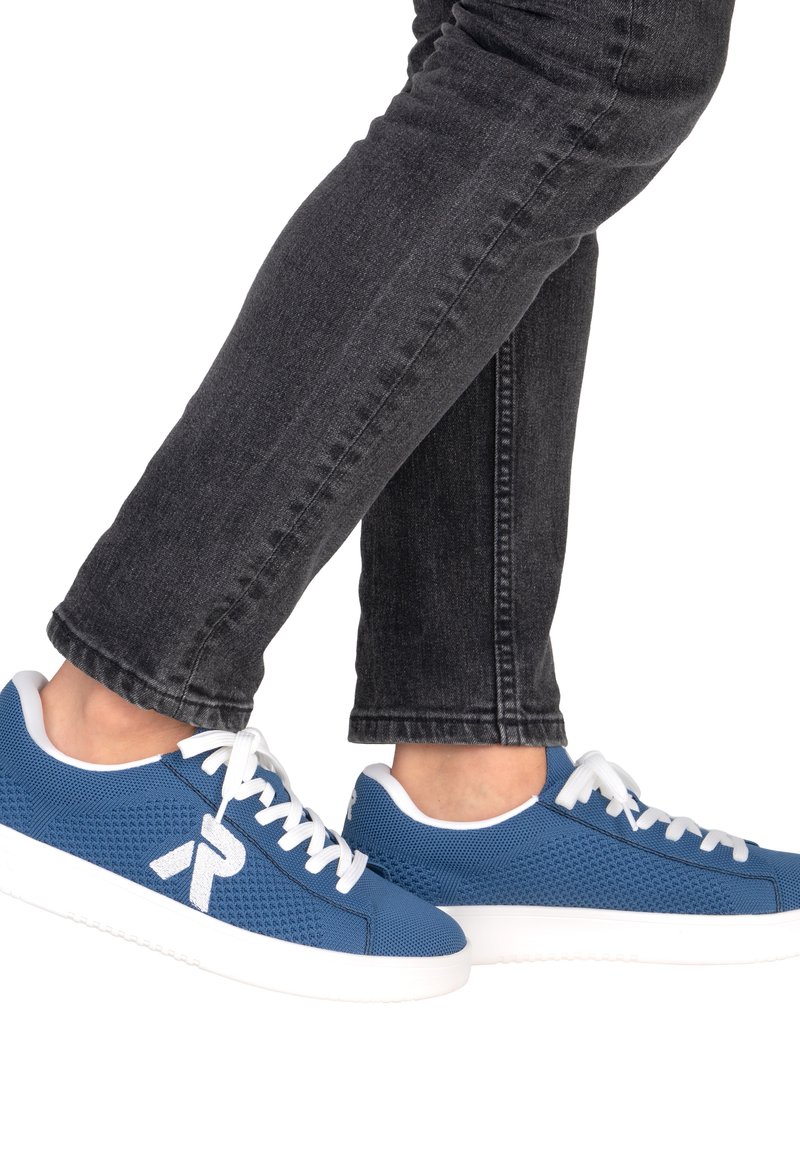Pieds portant des baskets en mesh bleu avec des lacets et semelles blanches, assorties à un jean en denim noir sur fond blanc.