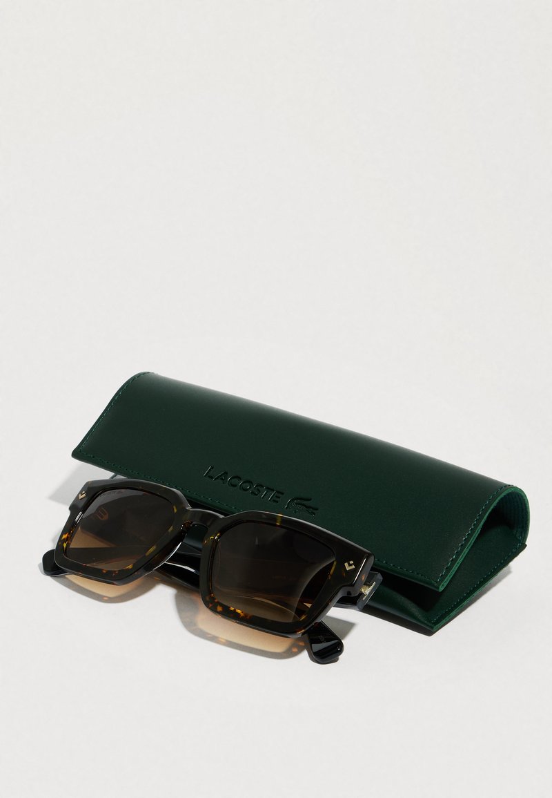 Skildpaddebrune solbriller med brune gradientlinser, der ligger på et mørkegrønt Lacoste-etui på en hvid overflade.