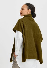 Poncho lavorato a maglia verde oliva con collo alto, caratterizzato da una vestibilità morbida e bordi grezzi, indossato sopra una camicia bianca e pantaloni chiari.