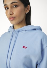 Felpa leggera blu chiaro realizzata in tessuto morbido con cappuccio regolabile. Presenta un piccolo logo rosso di Levi's sul davanti. Texture liscia e design casual.