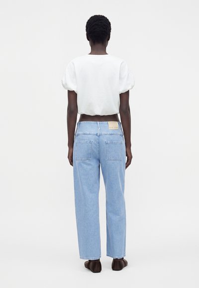 Personne aux cheveux courts et bouclés portant un haut blanc à manches courtes et un jean bleu clair à jambes droites, debout de dos sur un fond uni.