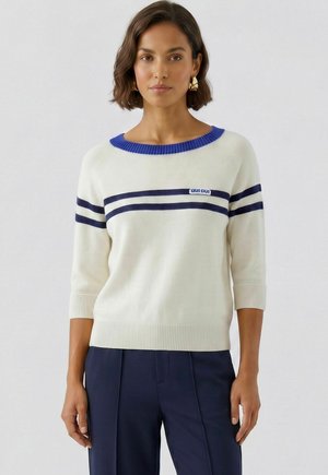 Vrouw met kort bruin haar, die een witte trui met blauwe strepen en kraag draagt, en donkerblauwe broek, staand tegen een effen achtergrond.