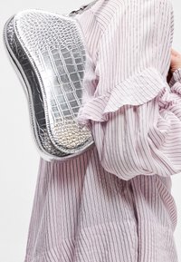 Personne portant un chemisier rayé rose et blanc tenant un sac à main argenté texturé brillant sur son épaule.