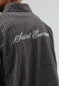 Bruin geruit overhemd met blauwe accenten, voorzien van een gekromde kraag en geborduurde tekst "Saint Essence" op de achterkant. Gestructureerde stof.