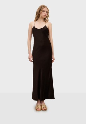 Joven mujer vistiendo un vestido largo negro tipo slip con finas tiras de cadena dorada y sandalias doradas de punta abierta, de pie frente a un fondo liso.