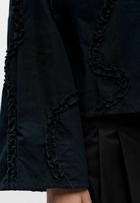 Czarna kurtka z falbankami wzdłuż rękawów. Tkanina wydaje się gładka, z wyraźnie zarysowanymi elementami konstrukcyjnymi. Subtelne plisowanie na dole.