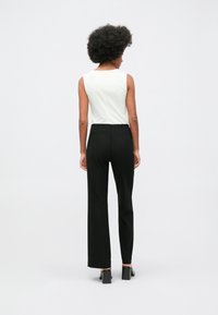 Mulher com cabelo afro de pé, de costas, vestindo uma blusa branca sem mangas, calças pretas e sapatos de salto bloco pretos.