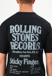 Widok z tyłu osoby w czarnej koszulce z jasnoszarym napisem promującym wytwórnię Rolling Stones Records oraz album Sticky Fingers z wymienionymi tytułami piosenek.